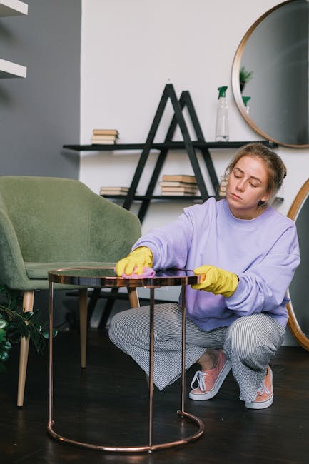 A person wearing black gloves is using a handheld electric upholstery steamer to deep clean a dark gray fabric sofa in a domestic living room setting. The sofa has a clean, well-maintained appearance