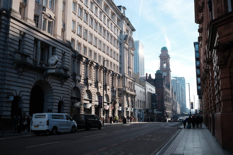Street scene depicting a busy urban area with classic and modern buildings under a clear blue sky. The foreground shows a wide sidewalk with pedestrians and a white van parked along the curb, while th