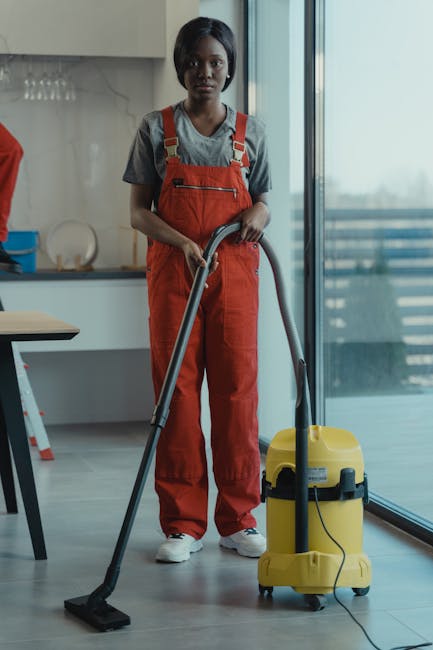 A professional domestic cleaner of African descent, wearing a grey T-shirt and orange overalls, is standing on a modern tiled floor with a vacuum cleaner in hand. The cleaner is positioned in a well-l