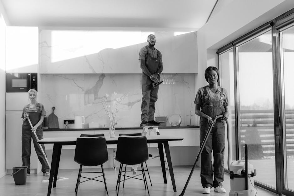 A professional domestic cleaner of African descent, wearing a grey T-shirt and orange overalls, is standing on a modern tiled floor with a vacuum cleaner in hand. The cleaner is positioned in a well-l